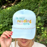 A person wearing The Darling Effect’s Dainty Pearl-Gold Trucker Hat Chain, styled on a light blue hat adorned with pearls and colorful “in my reading era” text, stands before a backdrop of blurred greenery. They hold the brim of the hat, displaying red-painted nails.