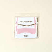 The Darling Effect Crystal Gem Trucker Hat Chain features metallic chains and rhinestones, packaged on a card with a pink hat outline and the brand’s name.