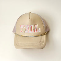 The Darling Effect’s beige trucker hat features pink and white embroidered "YALL" on the front and a Crystal Gem Trucker Hat Chain as a decorative gold detail, all set against an off-white background.