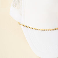 A white baseball cap with a white mesh back, featuring The Darling Effect's Crystal Gem Trucker Hat Chain in gold and rhinestones along the bill’s edge, shown against a light beige background.
