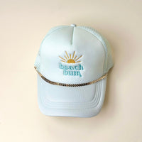A light blue trucker hat with "beach bum" and a yellow sun embroidered on the front features The Darling Effect's Gold-Leaf Chain Trucker Hat Chain above the brim, displayed on a beige background.