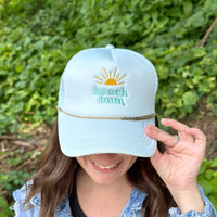 A woman smiles, holding the brim of her light blue "beach bum" cap with a sun graphic, styled with The Darling Effect's Gold-Leaf Chain Trucker Hat Chain, standing before lush greenery.