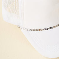 A white mesh trucker hat featuring The Darling Effect's Silver-Leaf Chain Trucker Hat Chain as a bold front-brim accessory, photographed on a light beige background.