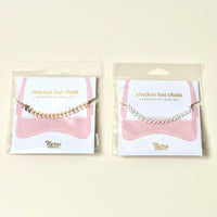 Two Silver-Leaf Chain Trucker Hat Chains by The Darling Effect are packaged on cards with pink wavy lines and the brand logo. One accessory is gold, the other silver—both add flair to mesh-back caps with chic chevron designs.