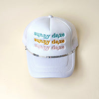 A white trucker hat with mesh back features "sunny daze" embroidered in blue, yellow, and peach. The Darling Effect's Silver-Leaf Chain Trucker Hat Chain decorates the front above the brim. Background is off-white.
