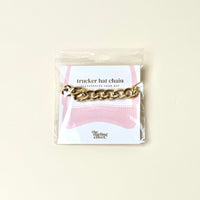 The Darling Effect Acrylic Link Gold Trucker Hat Chain shines on a light background and is packaged on a card with pink hat graphics and "the darling effect" at the bottom.