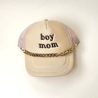 A beige trucker hat features "boy mom" embroidered in dark brown and is styled with The Darling Effect's Acrylic Link Gold Trucker Hat Chain, a bold accessory draped across the brim.