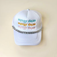 A white baseball cap with sunny daze embroidered three times in pastel blue, yellow, and orange, featuring a metallic chain draped across the front above the brim for a bold hat accessory vibe.