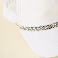 Close-up of The Darling Effect’s Acrylic Link Silver Trucker Hat Chain accenting a white mesh-back cap with a shiny silver link, shown on a light surface.