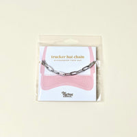 The Darling Effect Paperclip Chain-Silver Trucker Hat Chain is the perfect hat accessory, packaged on a card with a pink hat graphic and metallic chains. Text: trucker hat chain – accessorize your hat. The Darling Effect logo appears at the bottom.