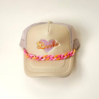 The Darling Effect's beige mesh trucker hat features a heart and "Books" embroidered on the front, accented with an Acrylic Sassy Trucker Hat Chain in pink and orange across the brim. The hat is shown on a plain white background.