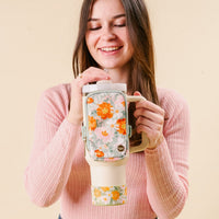 A smiling woman in a pink sweater holds the-darling-effect’s Bouquet Beauty Sage Tumbler with a colorful floral pattern, matching pouch, and protective silicone boot, set against a beige background.