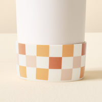 A close-up of The Darling Effect’s Check Brown Tumbler Silicone Boot—a white cylindrical container with a ceramic base in orange, brown, beige, and white checkered pattern—shown with its protective silicone boot.
