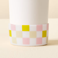 A close-up of The Darling Effect's Check Green Tumbler Silicone Boot, a white cylindrical tumbler accessory with a checkered silicone base in green, pink, yellow, beige, and white for added protection on a neutral surface.