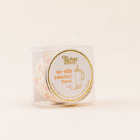 A clear plastic box holds the-darling-effect’s Endless Daydream Cream Tumbler Silicone Boot—a floral-patterned protector that prevents tumbler dents—with gold and orange text on a circular front label.