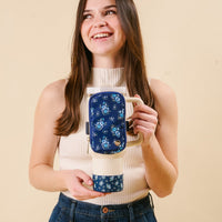 A smiling woman in a sleeveless white top holds the-darling-effect's Endless Daydream Navy Tumbler with a blue floral pouch and protective silicone boot, standing against a plain beige background.