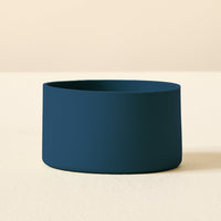 The Darling Effect's Navy Tumbler Silicone Boot, a dark blue, cylindrical accessory with smooth sides and a no-slip base for tumbler protection, rests on a light textured surface against a beige background.
