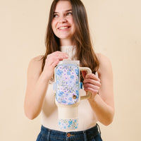 A smiling woman with long brown hair holds a cream-colored cup featuring the-darling-effect Secret Garden Blue Tumbler Silicone Boot and matching floral pouch, both offering stylish protection against dents, set against a beige background.