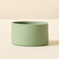The Darling Effect's Sage Tumbler Silicone Boot is a sleek, matte light green cylindrical accessory with smooth sides and a no-slip base, shown on a lightly textured surface against a beige background.