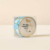 A clear plastic box showcases the Sweet Meadow Green Blue Tumbler Silicone Boot by The Darling Effect, featuring blue floral patterns and gold text on the lid for stylish, no-slip tumbler protection.
