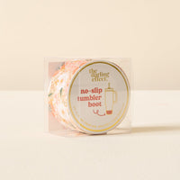 The Darling Effect’s Sweet Meadow Orange Tan Tumbler Silicone Boot is shown in a clear plastic box with a round front label against a light beige background—the perfect floral no-slip accessory for your tumbler.