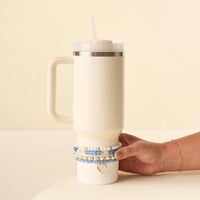 A hand holds a white 40 oz tumbler adorned with the-darling-effect's On Cloud Nine Tumbler Bracelet Stack, featuring stacked beaded bracelets and a heart-shaped charm.