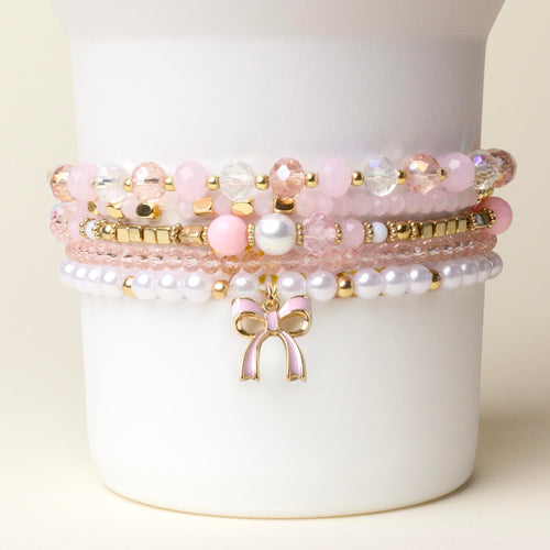 The-darling-effect Coquette Charm Tumbler Bracelet Stack features pink, white, and gold beaded bracelets with a gold bow charm—perfect as chic tumbler accessories or a stylish bracelet stack.