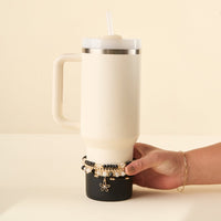 A hand holds a cream 40 oz tumbler with a straw lid, resting in a black holder adorned with the-darling-effect’s Midnight Glam Tumbler Bracelet Stack—black, white, and gold beads plus a gold flower charm.