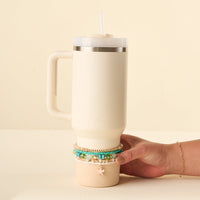 A hand adorned with the Tidal Wave Tumbler Bracelet Stack by the-darling-effect—featuring multiple colorful beaded bracelets and a starfish charm—holds a large, cream-colored 40 oz tumbler with a handle and straw.