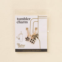 The Checkmate Tumbler Charm by the-darling-effect features a gold chain with three charms—a black flower, a black-and-white checkered heart with a bow, and a small star—perfect for personalizing your tumbler.