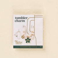 The Enchanted Tumbler Charm by the-darling-effect features a beaded chain with gold and green flower, gold flower, and pink heart charms. The packaging reads "Tumbler Charms" and "the darling effect." Accessorize your tumbler in style!.