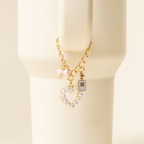The Glamour Tumbler Charm by the-darling-effect features a gold chain bracelet with three charms—a pink bow, black perfume bottle, and pearl heart—on a cream stand, perfect for pairing with your favorite tumbler accessories.