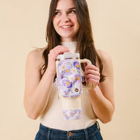 A smiling woman with long brown hair holds a beige mug and wears the-darling-effect's Bouquet Beauty Purple Tumbler Fanny Pack, featuring a stylish floral design. She's dressed in a sleeveless beige top and blue jeans against a plain backdrop.