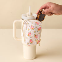 A hand places sunglasses into the-darling-effect’s Endless Daydream Cream Tumbler Fanny Pack, a stylish, water-resistant floral pouch attached to a cream tumbler with straw, resting on a light, textured surface.