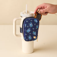 A hand puts sunglasses into the-darling-effect Endless Daydream Navy Tumbler Fanny Pack, which is attached to the handle of a large white mug with a straw.