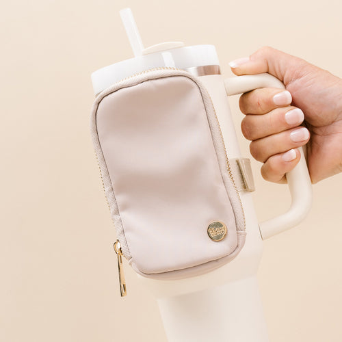 A hand holds The Darling Effect’s cream tumbler with a Natural Beige Tumbler Fanny Pack attached to its side and a white straw extending from the lid.