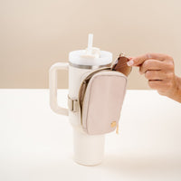 A hand places a key into The Darling Effect’s Natural Beige Tumbler Fanny Pack, attached to the handle of a large white tumbler with a straw on a light surface against a plain background.