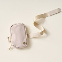 The Darling Effect’s Natural Beige Tumbler Fanny Pack—water resistant with a gold zipper, matching adjustable strap, and a round metallic emblem—lies flat on a light textured surface, showcasing its small and stylish design.