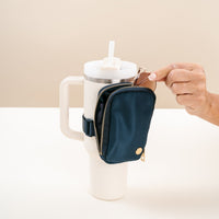 A hand unzips The Darling Effect Navy Tumbler Fanny Pack, attached to the handle of a large white travel tumbler with a straw lid, placed on a white surface with a beige background.