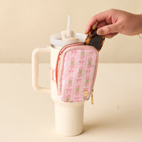 A hand places sunglasses into the-darling-effect's Oh So Fancy Pink Tumbler Fanny Pack, attached to a large cream tumbler with handle and straw, set on a beige surface.