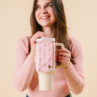 A young woman with brown hair smiles while holding the-darling-effect Oh So Fancy Pink Tumbler Fanny Pack, featuring a tan tumbler with a pink floral sleeve, and wears a stylish pink ribbed sweater.