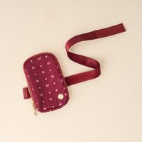 The Darling Effect's Pretty Posies Plum Tumbler Fanny Pack features a maroon pouch with a gold zipper, white floral pattern, and adjustable strap—ideal for carrying daily essentials in water resistant style.