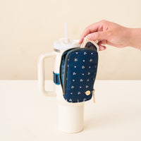 A hand places The Darling Effect Pretty Posies Teal Tumbler Fanny Pack, featuring a small floral pattern, onto the handle of a large white tumbler cup with a straw. The cup sits on a light surface against a beige background.