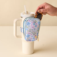 A hand places sunglasses into the-darling-effect's Secret Garden Blue Tumbler Fanny Pack, attached to a large white tumbler with a lid and two straws.