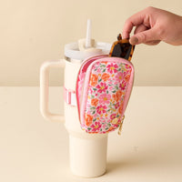 A hand places sunglasses into the-darling-effect's Secret Garden Pink Tumbler Fanny Pack, a floral-patterned, water-resistant pouch attached to a cream tumbler with a pink strap.