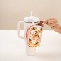 A hand opens The Darling Effect’s Sweet Meadow Orange Tan Tumbler Fanny Pack, a stylish floral zip pouch attached to the handle of a large white tumbler with straw; set against a neutral background on a white surface.