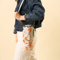 Wearing a denim jacket and cream pants, a person carries a mint green tumbler in the-darling-effect’s Bouquet Beauty Sage Tumbler Sling, featuring an adjustable strap for hands-free style.