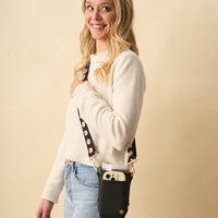 A smiling woman with long blonde hair wears a cream sweater and light blue jeans, carrying The Darling Effect's Black Tumbler Sling with a floral strap. A smartphone is partially visible inside.
