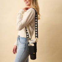 A woman in a white sweater and blue jeans stands sideways, smiling. She carries The Darling Effect’s Black Tumbler Sling, which has a floral strap and holds a water bottle and smartphone. The background is plain beige.