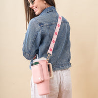A smiling woman in sunglasses and a denim jacket carries The Darling Effect’s Blush Tumbler Sling with a floral adjustable strap and phone pocket, perfectly complementing her white pants.